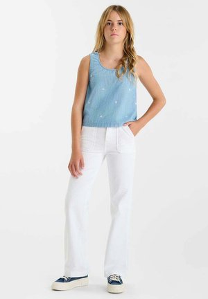 Tienermeisje staat met hand in de zak, draagt een lichtblauwe mouwloze top, witte wijde broekspijpen en marineblauwe sneakers, op een witte achtergrond.