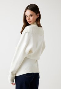 Biały, oversize'owy sweter z prążkowanym dekoltem typu off-shoulder i mankietami, noszony z ciemnymi dżinsami. Tekstura wydaje się miękka i przytulna.