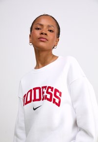 Valkoinen huppari, jossa on punaiset "GODDESS"-tekstit ja musta Nike-logo. Siinä on klassinen pyöreä kaula-aukko ja rento istuvuus. Sileä pinta.