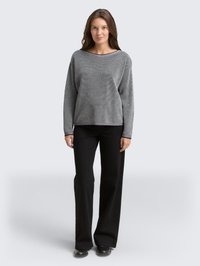 Donna in piedi di fronte, indossa un maglione grigio a maniche lunghe con texture, pantaloni neri a gamba larga e scarpe nere su uno sfondo neutro.