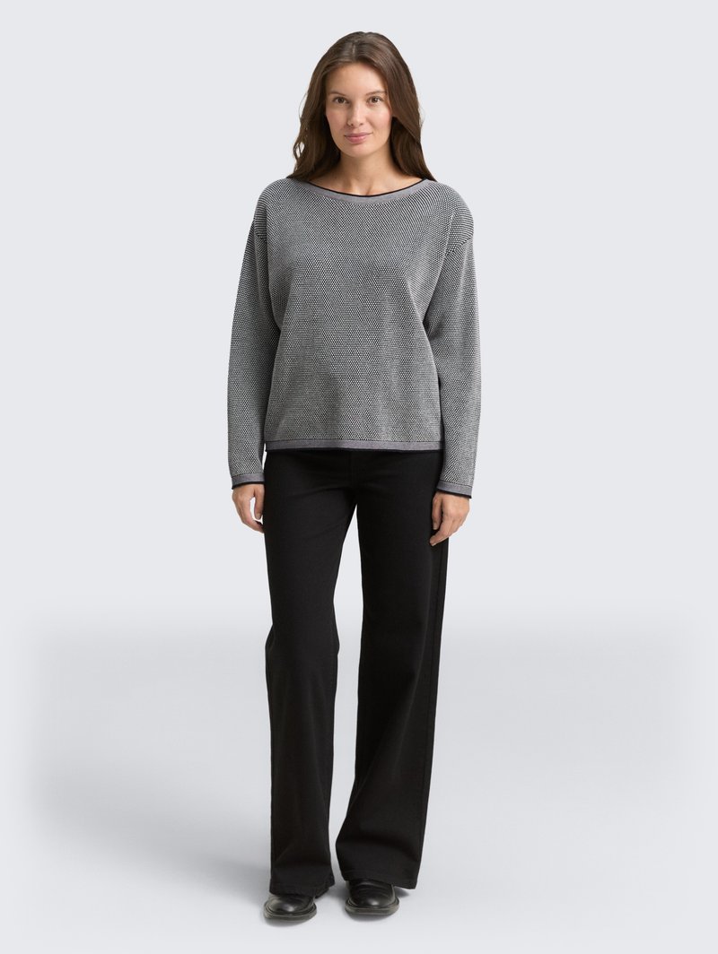 Kobieta stoi przodem, ubrana w szary sweter z długim rękawem o teksturowanym wykończeniu, czarne szerokie spodnie i czarne buty na jednolitym tle.