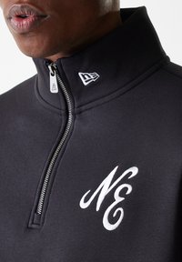 Schwarzer Fleece mit Reißverschluss und hohem Kragen, mit einem weißen gestickten "NE"-Logo auf der Brust und einem silbernen Reißverschluss-Zipper.