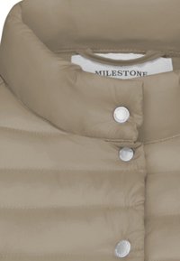 Beige Steppjacke mit hohem Kragen, silbernen Druckknöpfen, gestepptem Material und einem Etikett mit "MILESTONE" am Kragen.