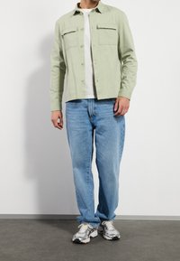 Hombre vestido con una chaqueta abotonada verde claro, camiseta blanca, vaqueros holgados azul claro y zapatillas blanco-gris, de pie contra una pared blanca.