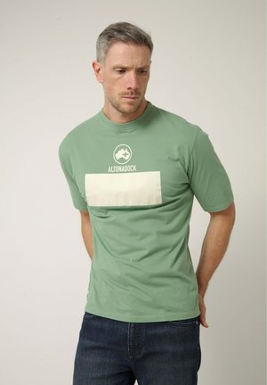 Hombre con pelo corto y gris, vistiendo una camiseta verde de Altonadock y vaqueros oscuros, mirando hacia abajo con las manos detrás de la espalda sobre fondo liso.