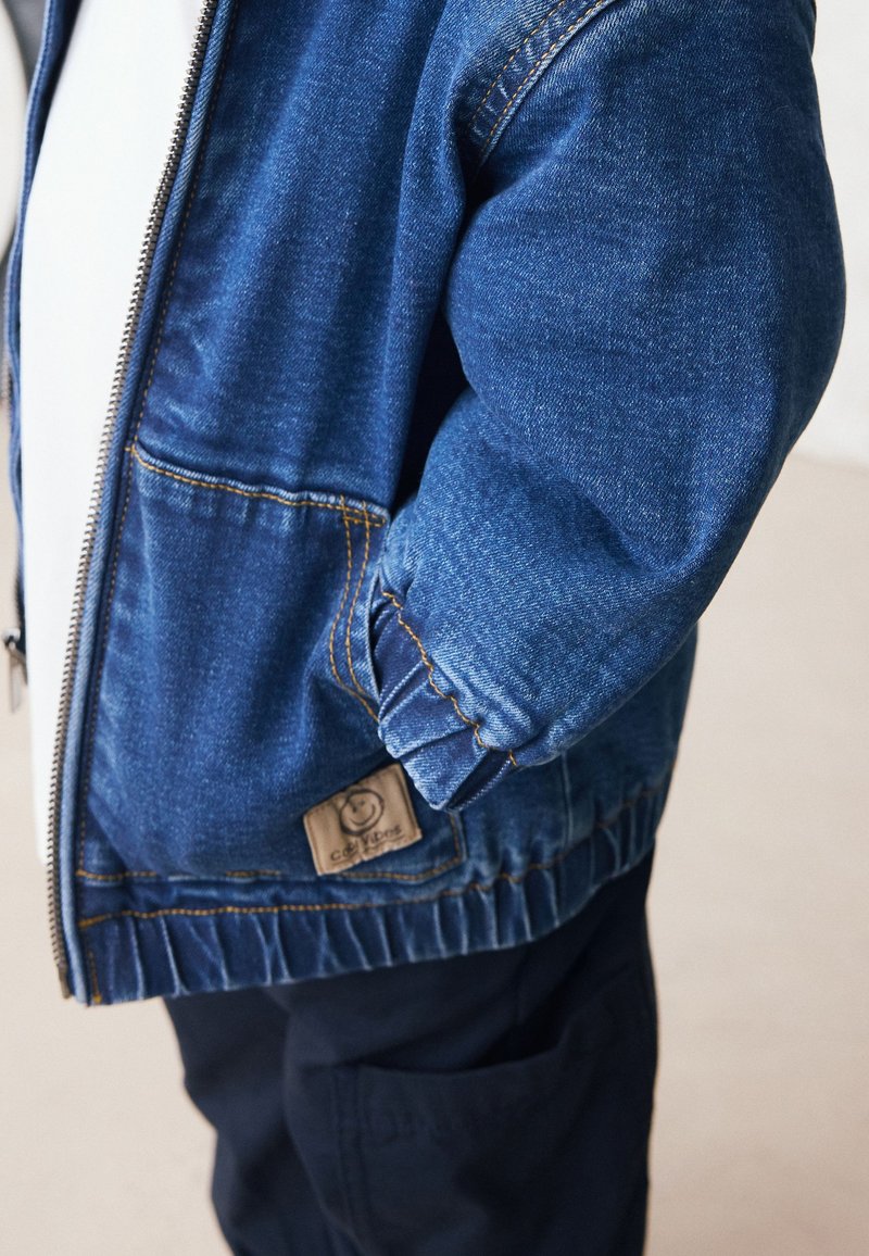 Bambino che indossa una giacca di denim blu con polsini elastici e tasche laterali, abbinata a pantaloni scuri, in piedi davanti a uno sfondo semplice.