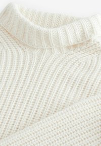 Gros plan d'un pull en maille blanche avec un col roulé côtelé épais et un motif texturé sur le corps et les manches.