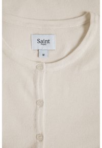 Krämfärgad cardigan med rund halsringning, utrustad med fem knappstängningar och en etikett med texten "Saint Tropez", storlek M. Mjuk textur.