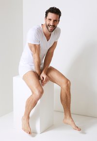 White cotton V-neck T-shirt and shorts, sitting on a white pedestal. Male model with short dark hair and lightly tanned skin. Natural posture.