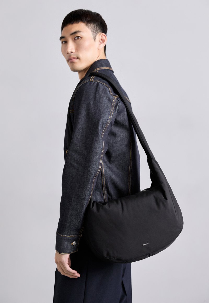 Borsa a spalla nera realizzata in tessuto morbido, caratterizzata da una forma curva, un design minimale e un piccolo logo del marchio sul fronte.