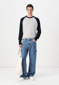 Grey and navy long-sleeve shirt with a heart design, paired with wide blue jeans. Model holds a beige tote bag and wears white sneakers.