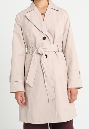 Beige knælang trenchcoat med bred krave, sorte knapper, bindebælte i taljen og justerbare håndledsremme båret af en person.
