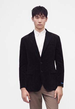 Jeune homme portant un blazer noir sur une chemise blanche boutonnée et un pantalon marron clair, debout devant un fond uni.