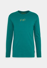 Long-sleeve teal t-shirt made of soft cotton. Features a round neckline and a gold cursive logo on the chest. Smooth texture, casual fit.