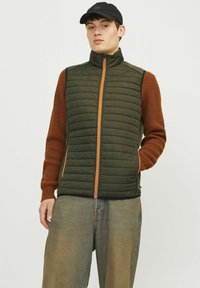 Vesta căptușită de culoare verde măslină cu fermoar portocaliu, purtată peste un pulover maro cu nervuri, asociată cu pantaloni din denim deschis.