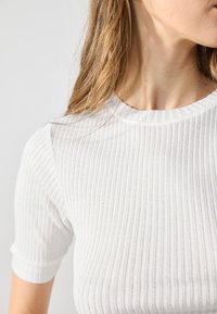 Top de punto acanalado blanco con mangas cortas, que presenta un escote redondo y una textura suave, resaltando las líneas verticales en la tela.