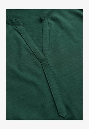 Dunkelgrüner Stoff mit V-Ausschnitt, der eine schmale, genähte Knopfleiste und eine dezente Textur aufweist.
