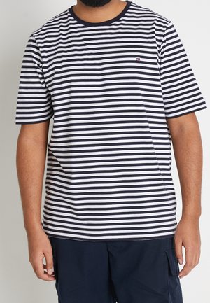 Man in een kortmouwen marineblauw en wit horizontaal gestreept T-shirt en donker marineblauwe broek, staande tegen een effen achtergrond.