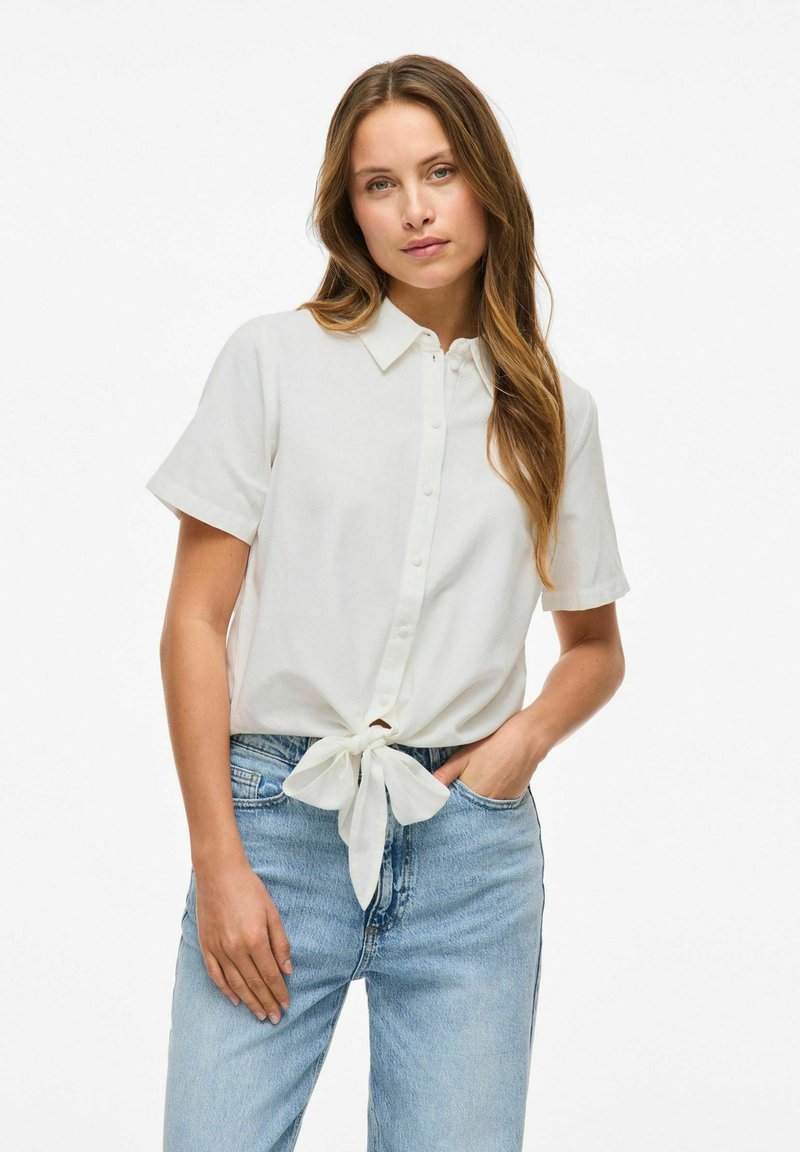 Camicia bianca a maniche corte con bottone, annodata in vita, abbinata a jeans blu chiaro. Tessuto morbido.