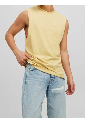 Mand iført ærmeløs lysegul skjorte med teksten "Originals" og lyseblå revnede jeans, der poserer foran en ensfarvet baggrund.