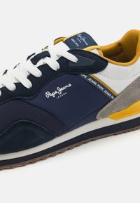 Sötétkék és fehér sneaker, velúrból és hálós anyagból készült. Sárga és szürke részletek, fehér fűzők és texturált gumitalp jellemzi.