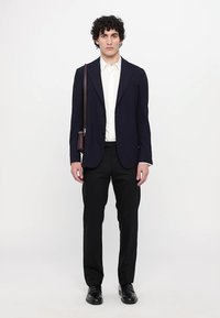 Blazer navy con rever a punte su una camicia bianca, abbinato a pantaloni neri. Inclusi una borsa a spalla marrone e scarpe nere lucide.