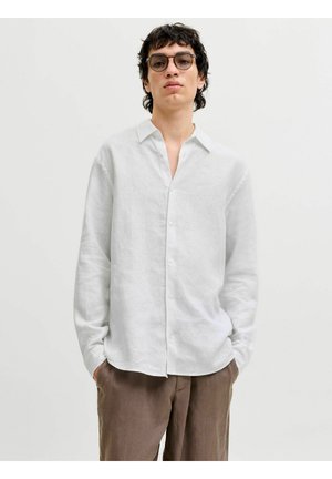 Jeune homme portant des lunettes rondes, une chemise blanche à boutons avec col, et un pantalon marron, debout les mains dans les poches devant un fond uni.