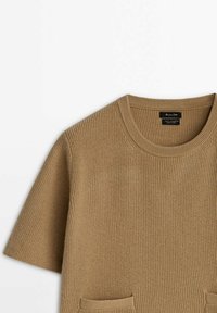 Pull en maille côtelée marron camel à manches courtes, col rond et petites poches avant sur fond blanc.