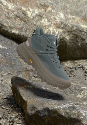 Γκρι πεζοπορικό μποτάκι Columbia Titanium με σόλα Vibram αιωρείται πάνω από πετρώδες έδαφος σε εξωτερικό χώρο.