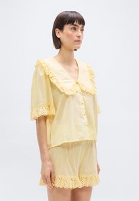 Femme portant un chemisier boutonné jaune clair et un short assorti avec des volants en dentelle, debout devant un fond blanc uni.