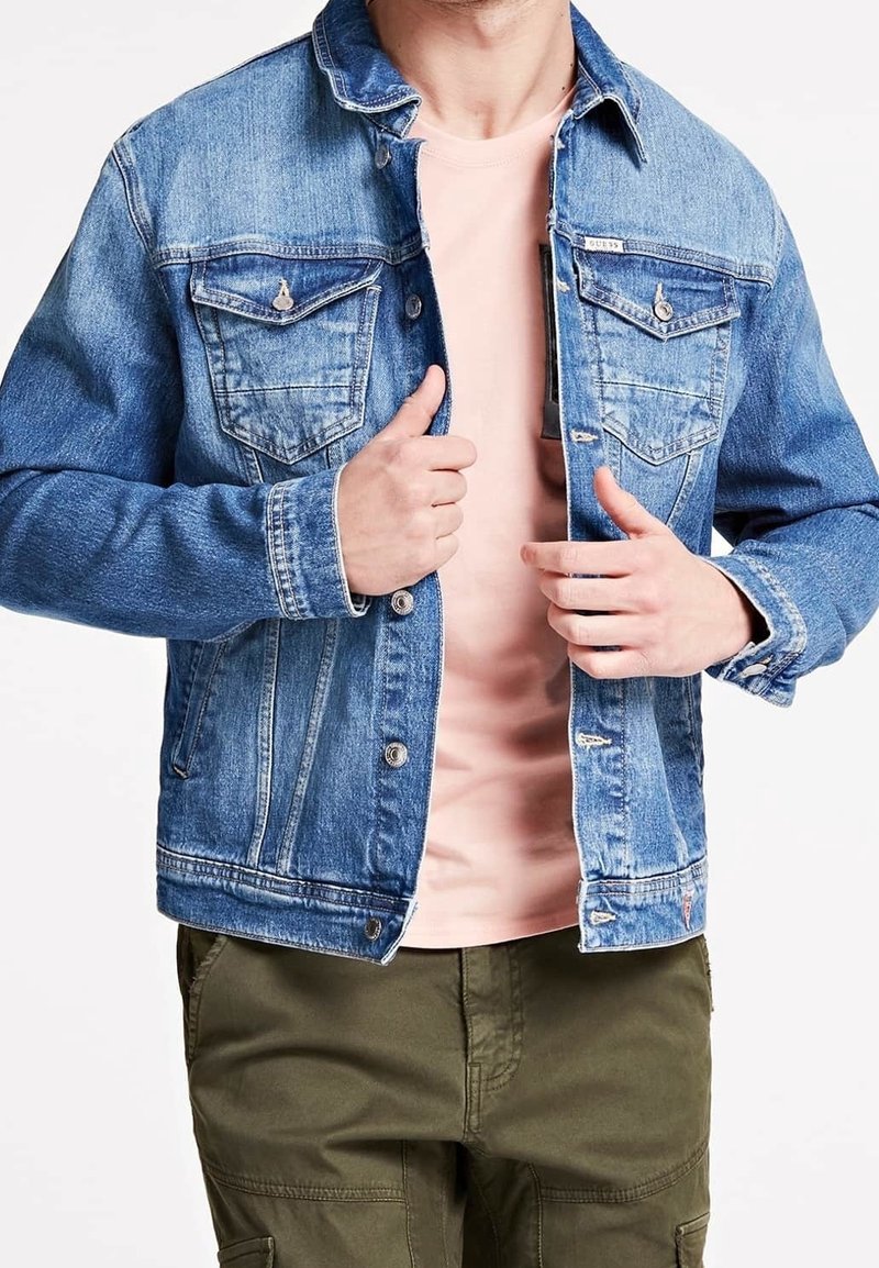 Vaaleansininen farkkutakki klassisella muotoilulla, jossa on kaksi rintataskua, napitus ja näkyvät ompeluyksityiskohdat. Tyylitelty vaaleanpunaisen paidan päällä.