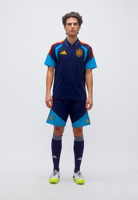 Jaunas vyras stovintis Ispanijos nacionalinės futbolo rinktinės uniformoje su tamsiai mėlynomis, žalsvai mėlynomis ir raudonomis detalėmis, dėvintis baltus sportinius batelius su neoninės geltonos spalvos pado dalimi.