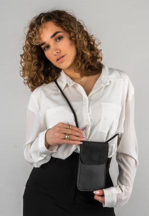 Sort læder crossbody taske med klap, båret på en sort snor. Båret over en hvid skjorte med knapper, der viser en glat tekstur og minimalistsk design.