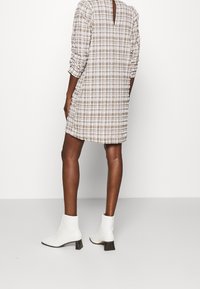 Brown and white checkered dress with long sleeves, a back slit, and gathered texture. Worn with white ankle boots featuring a block heel.