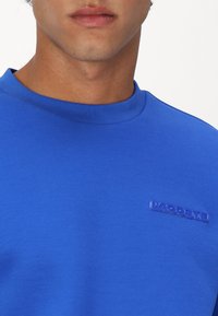 Sudadera azul texturizada con cuello redondo, que presenta un acento de logo en plástico elevado. La tela tiene un patrón de malla fina.