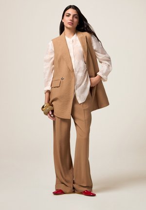 Beige Überwurfweste mit enganliegendem Oberteil, weitem Hosen in einem passenden Farbton und roten High Heels. Auffällige goldene Armreifen am Handgelenk.