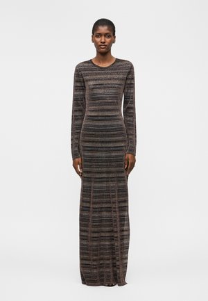 Femme aux cheveux courts portant une longue robe tricotée rayée marron et noire, ajustée, manches longues, debout devant un fond blanc uni.