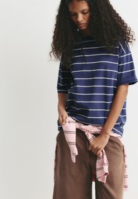 Marine-stribet t-shirt med hvide vandrette linjer, afslappet pasform. Brune bukser og en lyserød plaidskjorte bundet om taljen.