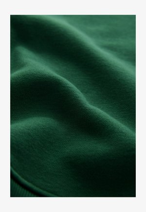 Tissu en coton vert, doux, présentant une texture lisse avec des variations subtiles de nuance et de légères aspérités, indiquant flexibilité et drapé.
