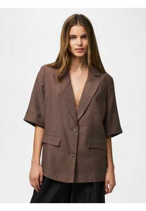 Žena nosiaca veľký hnedý blejzer s krátkymi rukávmi a dvoma prednými vreckami pózujúca na jednofarebnom bielom pozadí.