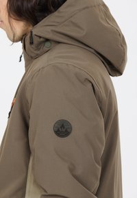 Brun huva jacka med en strukturerad yta, en rund svart patch på ärmen och justerbara element för passform och komfort.