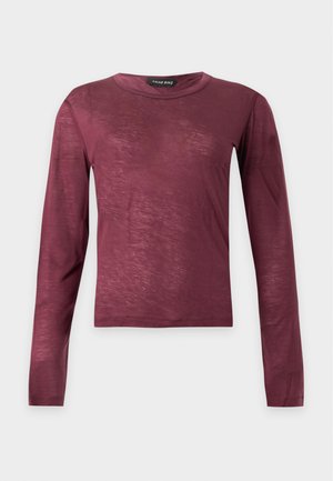 Vrouw met kort donker haar, een nauwsluitende bordeauxrode top met lange mouwen, wijde blauwe jeans en bruine puntige schoenen, staand in een neutrale houding.
