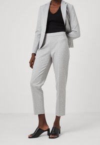 Kobieta w jasnoszarych, dopasowanych spodniach i marynarce, z czarnym bezrękawnikiem i czarnymi sandałami na obcasie z odkrytym palcem, stoi na jednolitym tle.