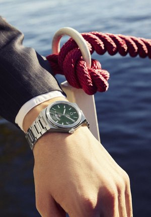 Mees ülikonnas hõbedase käekellaga, kelle käsi puhkab vee ääres punase nööri lähedal, mis on seotud valge metallrõngaga.