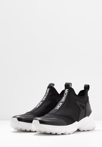 Czarne sneakersy na stopie z tekstylną cholewką, białą gumową podeszwą i wyraźnym logo na elastycznym pasku. Okrągły czubek i solidna konstrukcja.