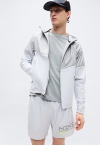 Αθλητικό μπουφάν σε ανοιχτό γκρι με φερμουάρ μπροστά και κουκούλα, συνδυασμένο με γκρι κοντομάνικη μπλούζα και ταιριαστό σορτς που φέρει το λογότυπο της Nike σε σκούρο γκρι με πράσινες λεπτομέρειες.