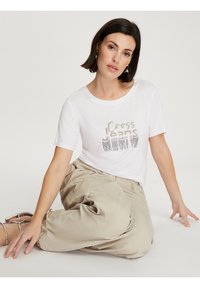 Biała koszulka z krótkim rękawem z nadrukiem w złotym i czarnym kolorze "Cross Jeans" oraz grafiką. Połączona z jasnobeżowymi, luźnymi spodniami.