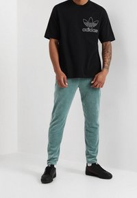 Černé tričko s krátkým rukávem s bílým logem Adidas; zelené velurové tepláky; černé tenisky. Jednoduchý design, měkké textury a kontrastní barvy.