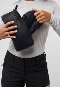 Zwarte pufferjack wordt in een klein pakket gestopt, vastgehouden door handen. De achtergrond is eenvoudig, en de drager draagt een lichtgekleurde top en donkere broek.