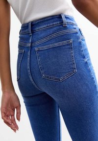 Vaqueros de mezclilla azul de cintura alta con una silueta ajustada. Las características incluyen bolsillos traseros, detalles de costura y una textura suave.